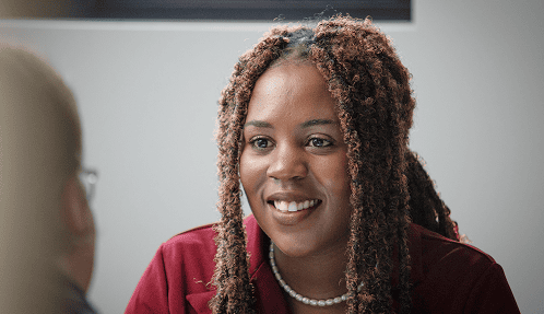 Woman with curly hair and a pearl necklace smiles while talking with someone opposite her.