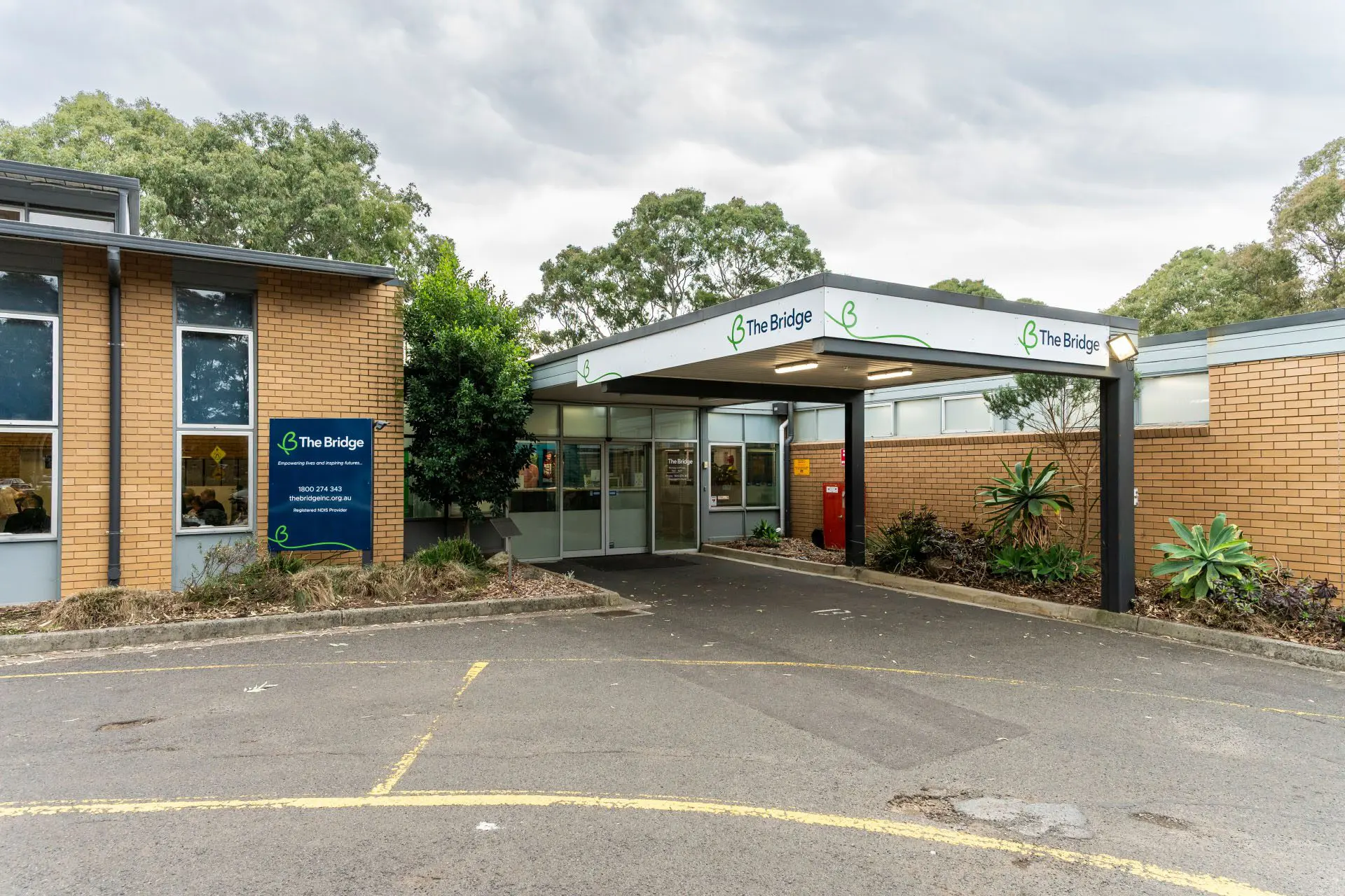 Brick building entrance with covered canopy and sign reading 'The Bridge', glass doors, and car park in front.