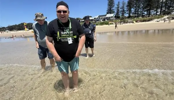 Three people wade in shallow sea water on a beach, smiling, with pine trees and buildings behind.