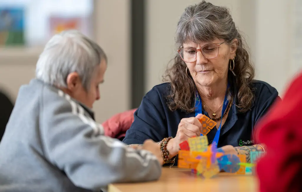 Staff member wearing a lanyard helps a person build with colourful construction toys at a table.
