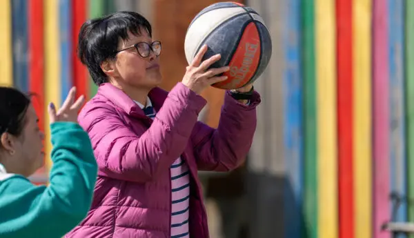 Woman in pink puffer jacket holds up a basketball to shoot, another person nearby, colourful striped wall behind.