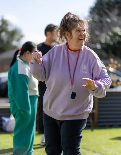 Smiling woman in a lavender sweatshirt and lanyard gestures outdoors, two people standing behind her on grass.