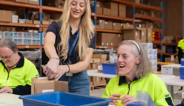Staff member with lanyard assists a smiling woman with a cardboard box at a warehouse workstation with blue bins.
