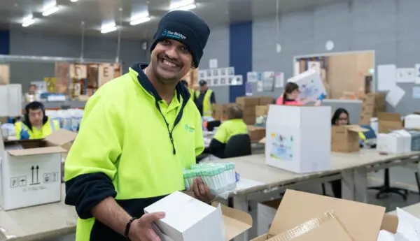 Man in hi-vis jacket and beanie smiling, holding boxed items, with colleagues packing boxes at tables behind.
