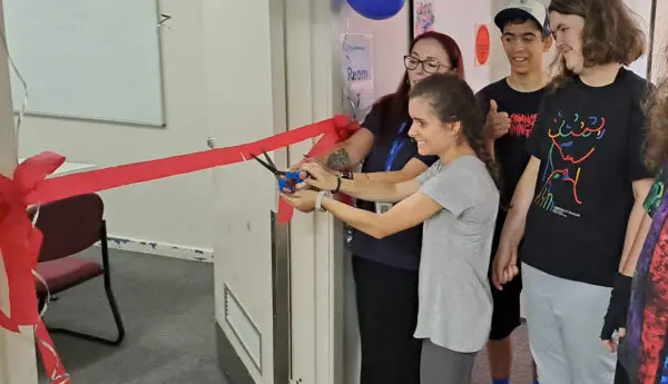 A woman cuts a red ribbon across a doorway with scissors, smiling, as others watch; blue balloons decorate the entrance.