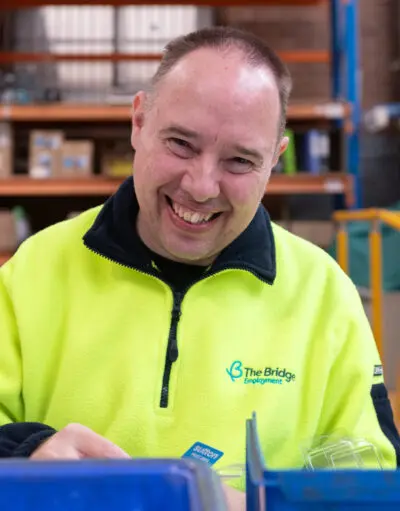 Smiling man in a hi-vis fleece with The Bridge Employment logo, working at a warehouse bench.
