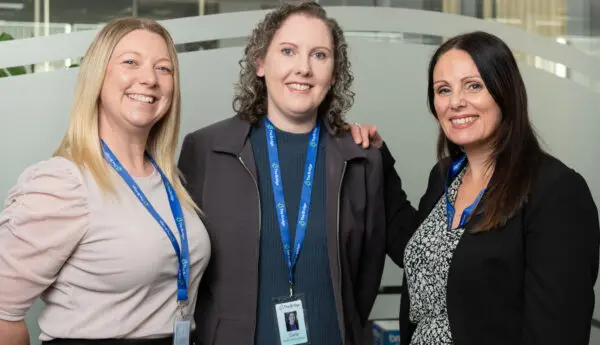 Three women wearing The Bridge lanyards stand smiling together in an office.