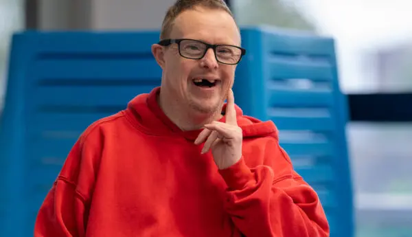 Man in a red hoodie and glasses smiles, raising a hand near his chin, seated in front of a blue chair.