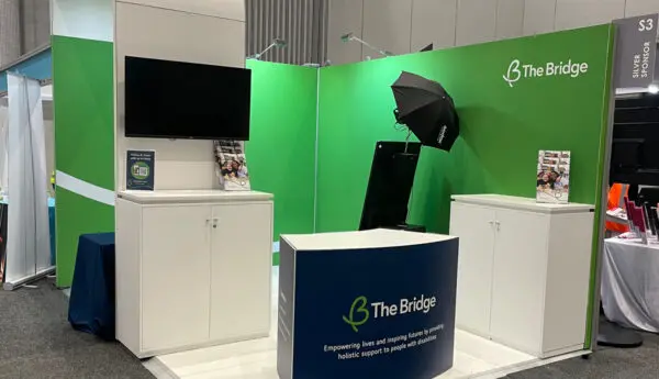 Green exhibition booth for The Bridge with a wall-mounted screen, photo booth with umbrella light, and navy counter sign.