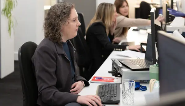Woman at a desk using a computer mouse, with two colleagues working at desks behind her in an office.