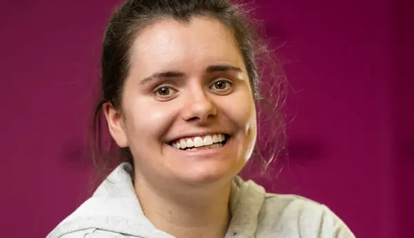 Woman with dark hair tied back, smiling at the camera, wearing a light grey hoodie against a maroon background.