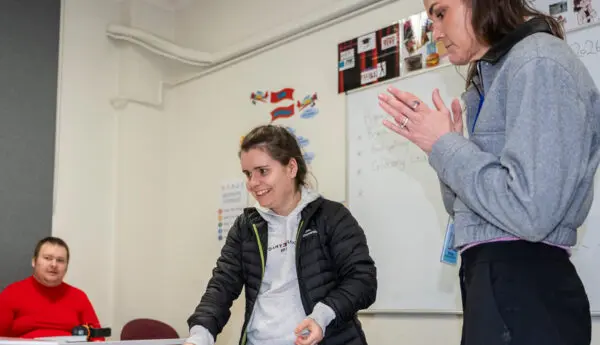 A woman smiles while placing cards on a table as a staff member gestures, with a man seated nearby.