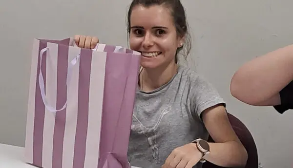 Woman smiling at a table, holding up a purple striped gift bag.