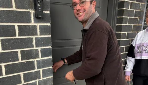 Man in glasses smiles while unlocking a grey front door, with a person standing behind him.