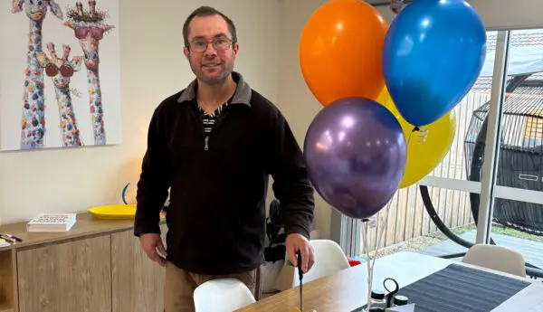 Man standing by a table with balloons, a cake, and a card reading Congratulations Matthew