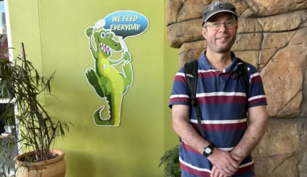 Man in a striped polo shirt stands by a wall with a crocodile cartoon reading 