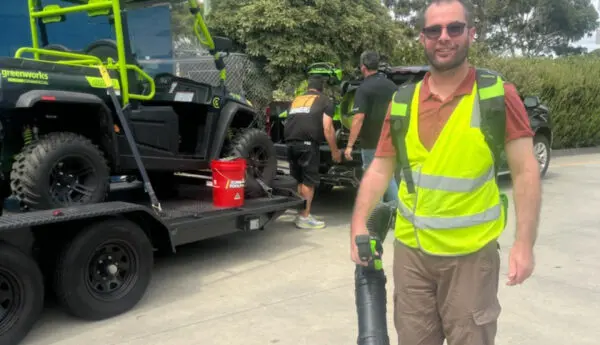 Man in hi-vis vest holds a leaf blower, smiling; behind him a utility vehicle on a trailer and two workers.