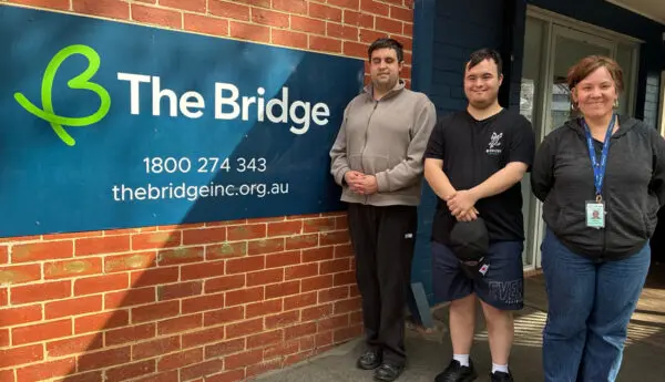 Three people stand beside a brick wall with The Bridge sign showing phone number and website.