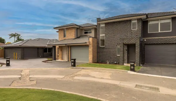 Row of two-storey brick townhouses with garages, driveways and letterboxes along a street.