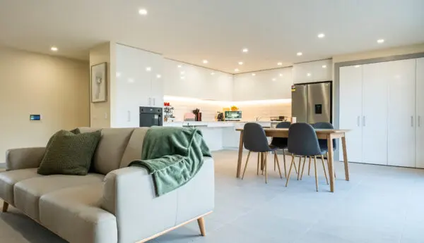 Open-plan living area with grey sofa, dining table and chairs, and white kitchen with stainless fridge.