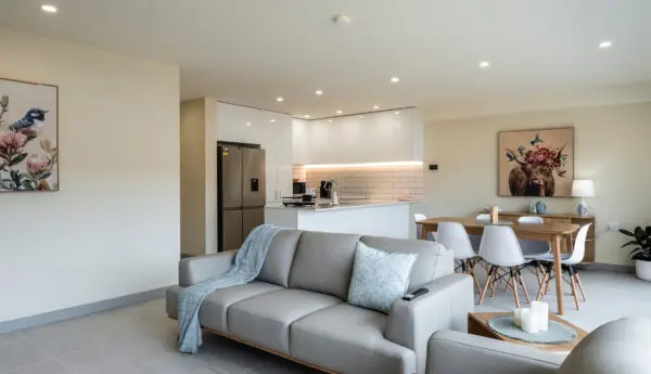 Open-plan living area with grey sofas, dining table, and white kitchen with stainless fridge.