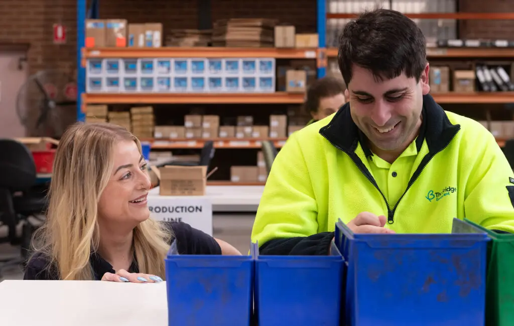 Man in high-vis jacket sorts items in bins at a table beside a smiling woman, in a warehouse.