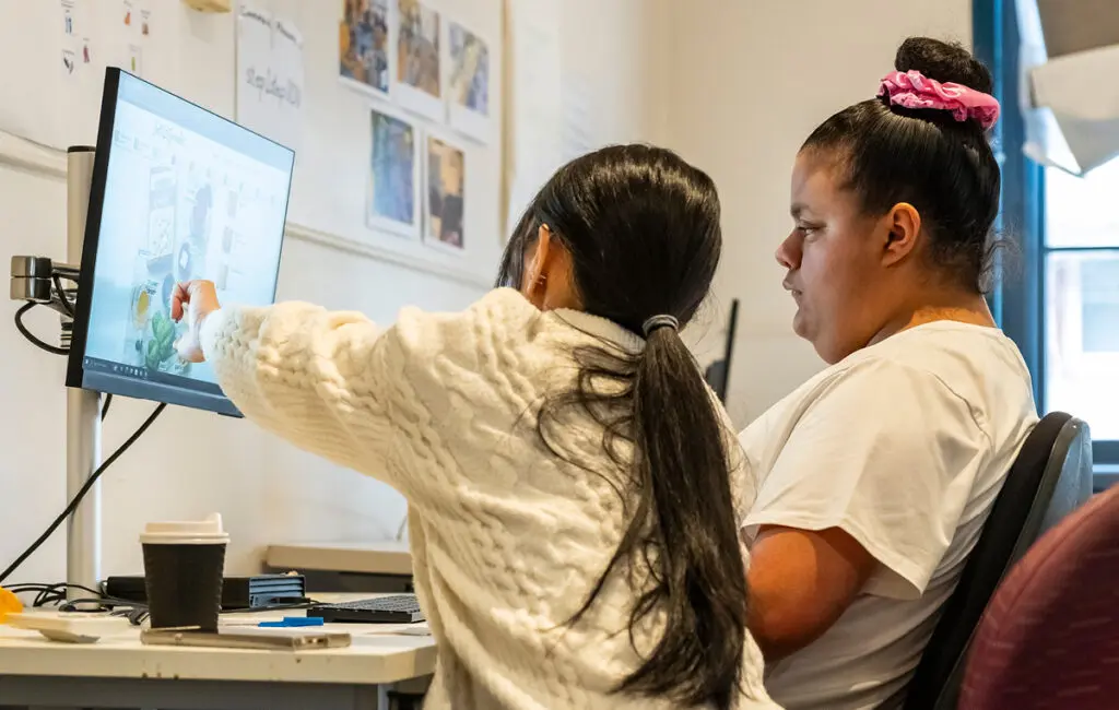 Two women look at a computer screen; one points at the display while the other sits and watches.