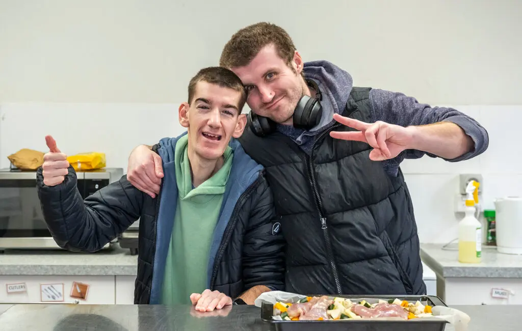 Two men smile in a kitchen, one giving a thumbs up, the other a peace sign, with a tray of raw food on the counter.