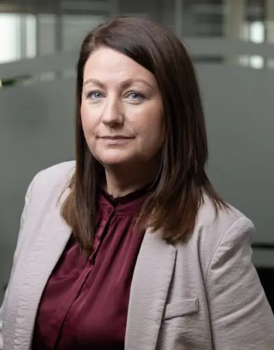 Woman with shoulder-length brown hair wears a beige blazer over a maroon blouse in an office setting.
