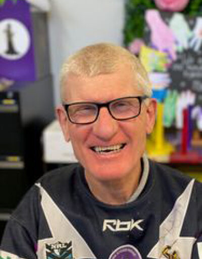 Man with glasses smiling in a black Reebok sports jersey, indoors with colourful craft displays behind him.