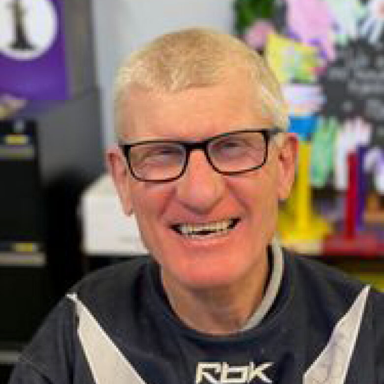 Man with glasses smiling in a black Reebok sports jersey, indoors with colourful craft displays behind him.