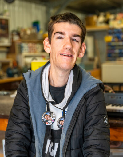 Man with badges on hoodie smiling at camera, standing in a workshop or shed.