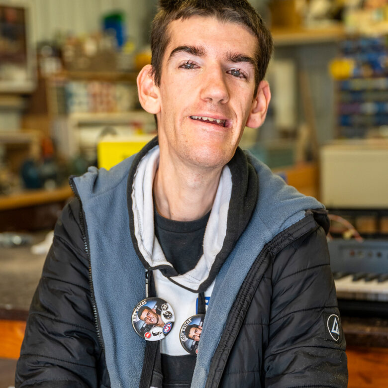 Man with badges on hoodie smiling at camera, standing in a workshop or shed.