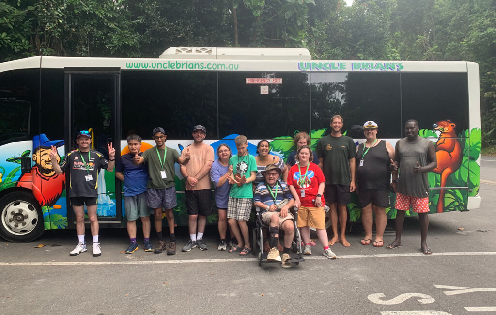 Group poses in front of an 'Uncle Brian's' tour bus, one person using a wheelchair, surrounded by rainforest.