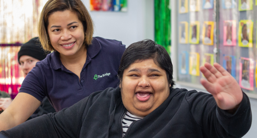 A woman raises her hand smiling, with a support worker in a 'The Bridge' polo shirt behind her in a classroom.