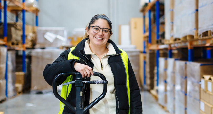 Woman in glasses and hi-vis jacket smiling, resting hand on pallet jack in a warehouse aisle.