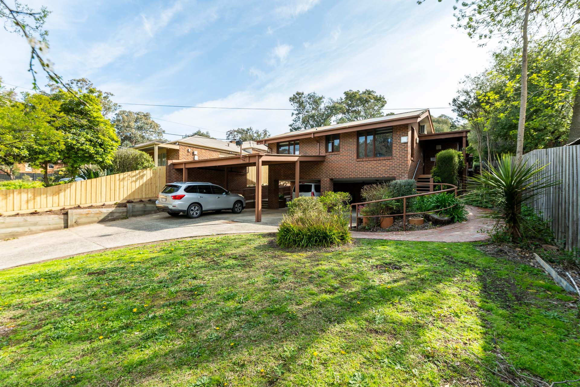 Brick house with carport, two parked cars, front lawn, garden beds and a curved brick path.